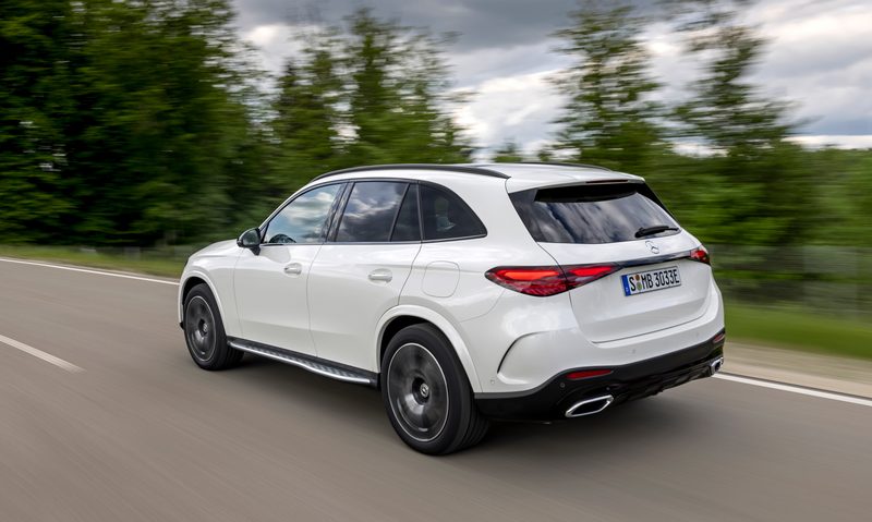 White 2025 Mercedes-Benz GLC 350e 4MATIC SUV driving with trees on the roadside left rear three-quarter view.