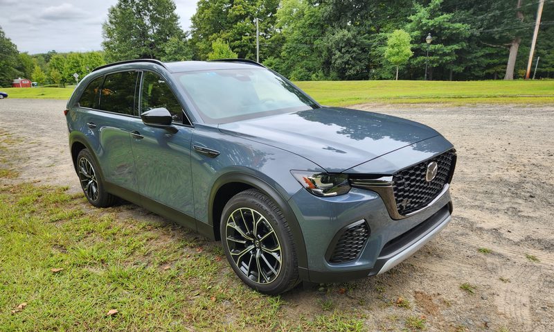2025 Mazda CX-70 PHEV right front three-quarter view parked in gravel lot surrounded by trees.
