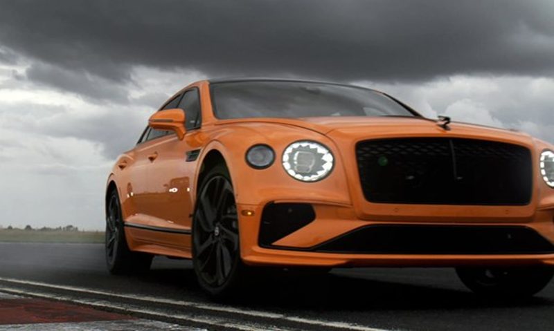 Bentley Flying Spur tease right front three-quarter view driving on a highway under a dark cloudy sky.