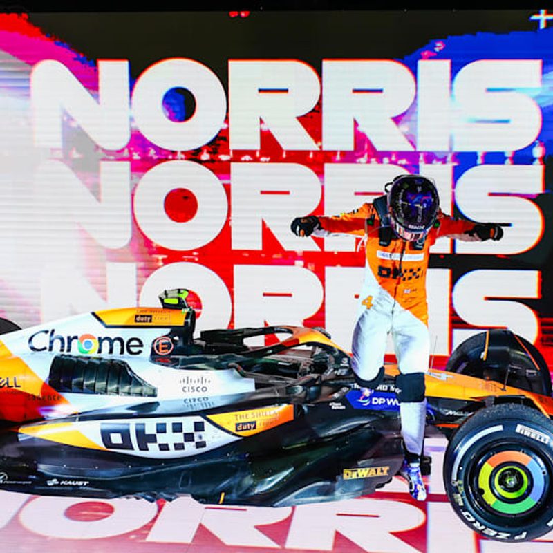 F1 driver Lando Norris standing victorious with his McLaren race car.