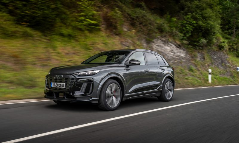 Gray Audi Q6 e-tron drivng on highway with forested hills in the background left front three-quarter view.