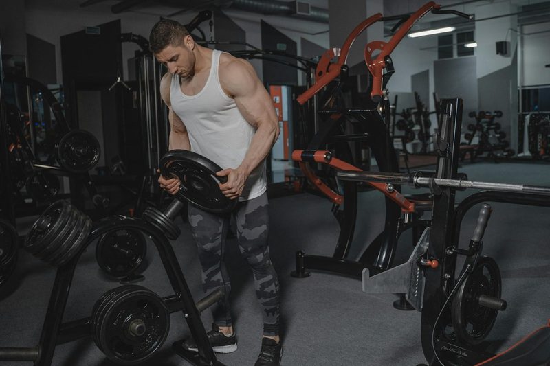 Man wearing a white tank top in the gym holding a weight and stacking weights on a rack