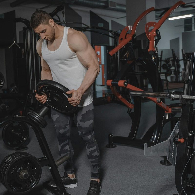 Man wearing a white tank top in the gym holding a weight and stacking weights on a rack