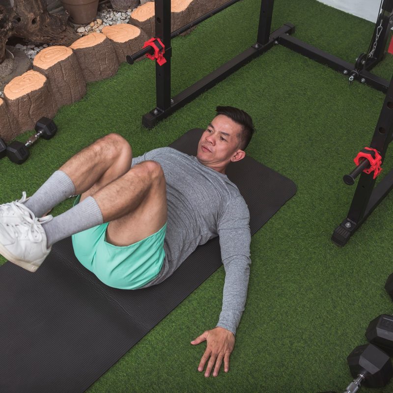 Man wearing long grey sleeve shirt with green shorts performing reverse crunch exercise on black exercise mat outside