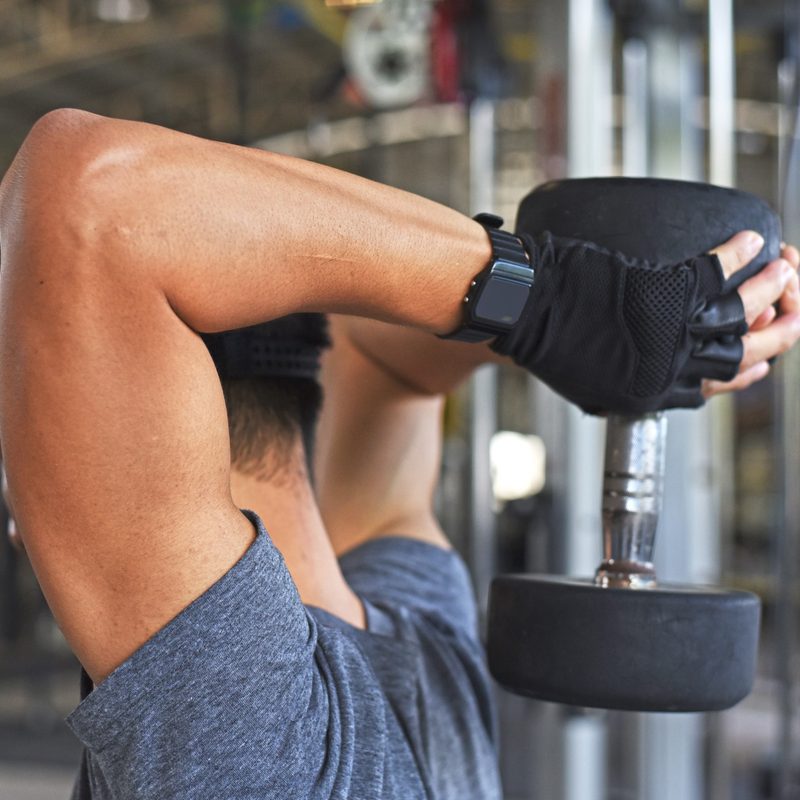 Man in black hat and blue T-shirt doing an overhead tricep extension with dumbbell in the gym
