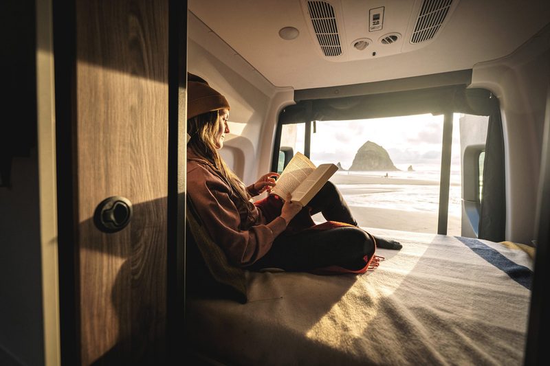Person reading in bed with a view of the ocean from inside a Tiffin GH1 camper van.