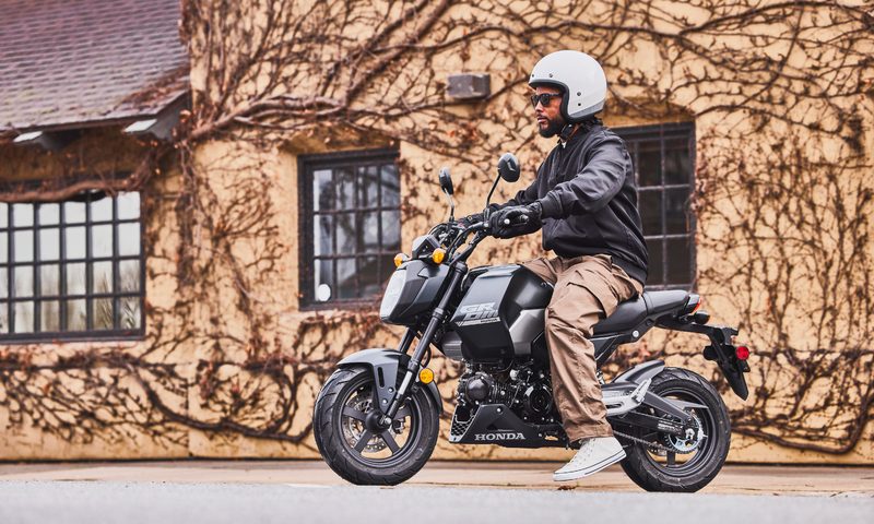 A person on a 2025 Honda Grom SP on a city street with a vine-covered stucco building in the background.