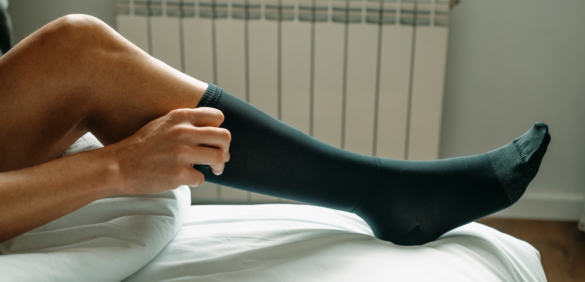 a man puts on a black compression sock sitting on his bed at home