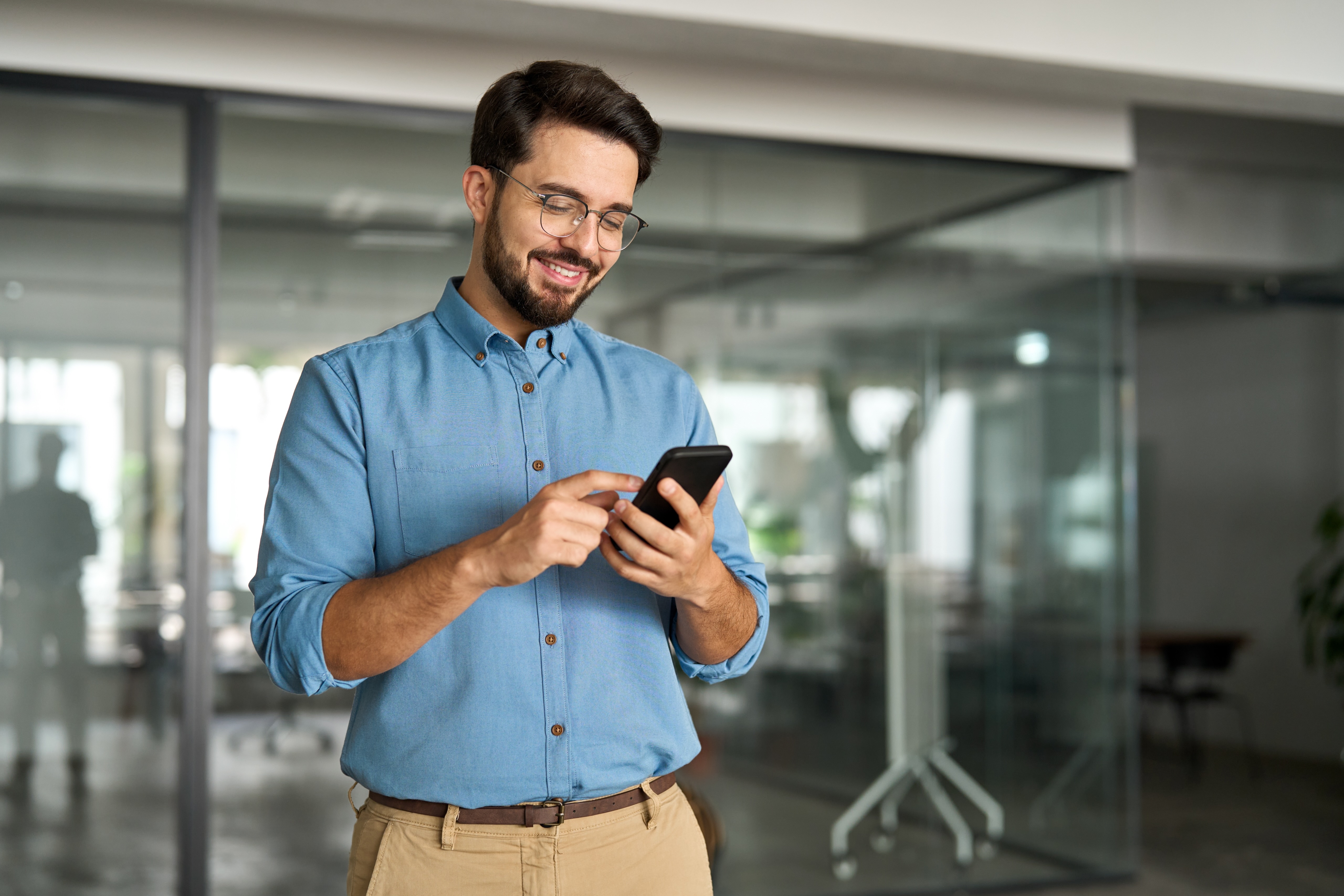 Happy young latin business man holding smartphone standing in office