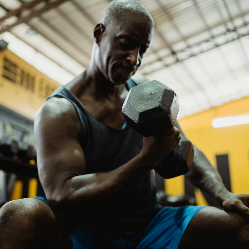Man wearing blue tank sitting in gym doing dumbbell concentration curl on bench