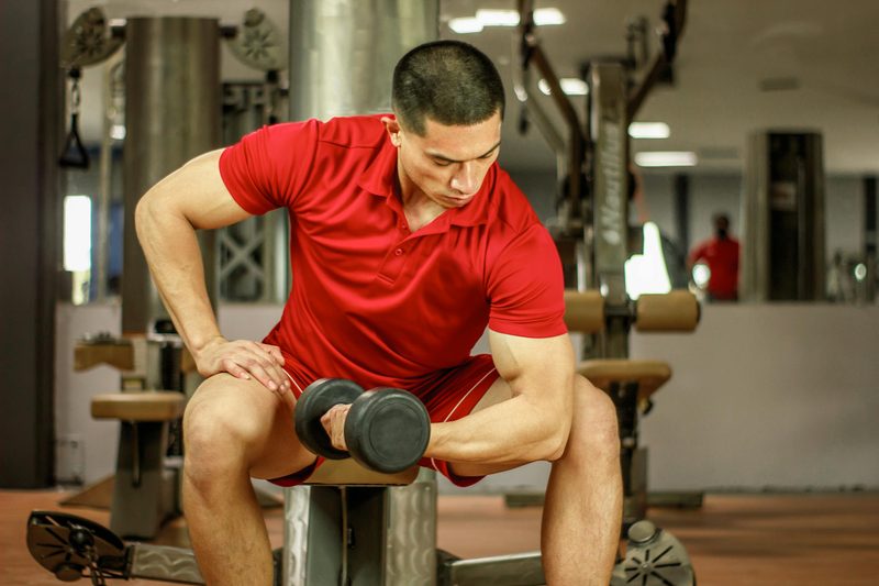 Man in gym wearing red shirt doing concentration curl with dumbbell seated on bench