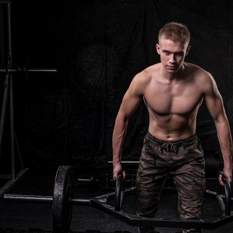 Shirtless man on a dark background performing trap bar deadlift with hex bar