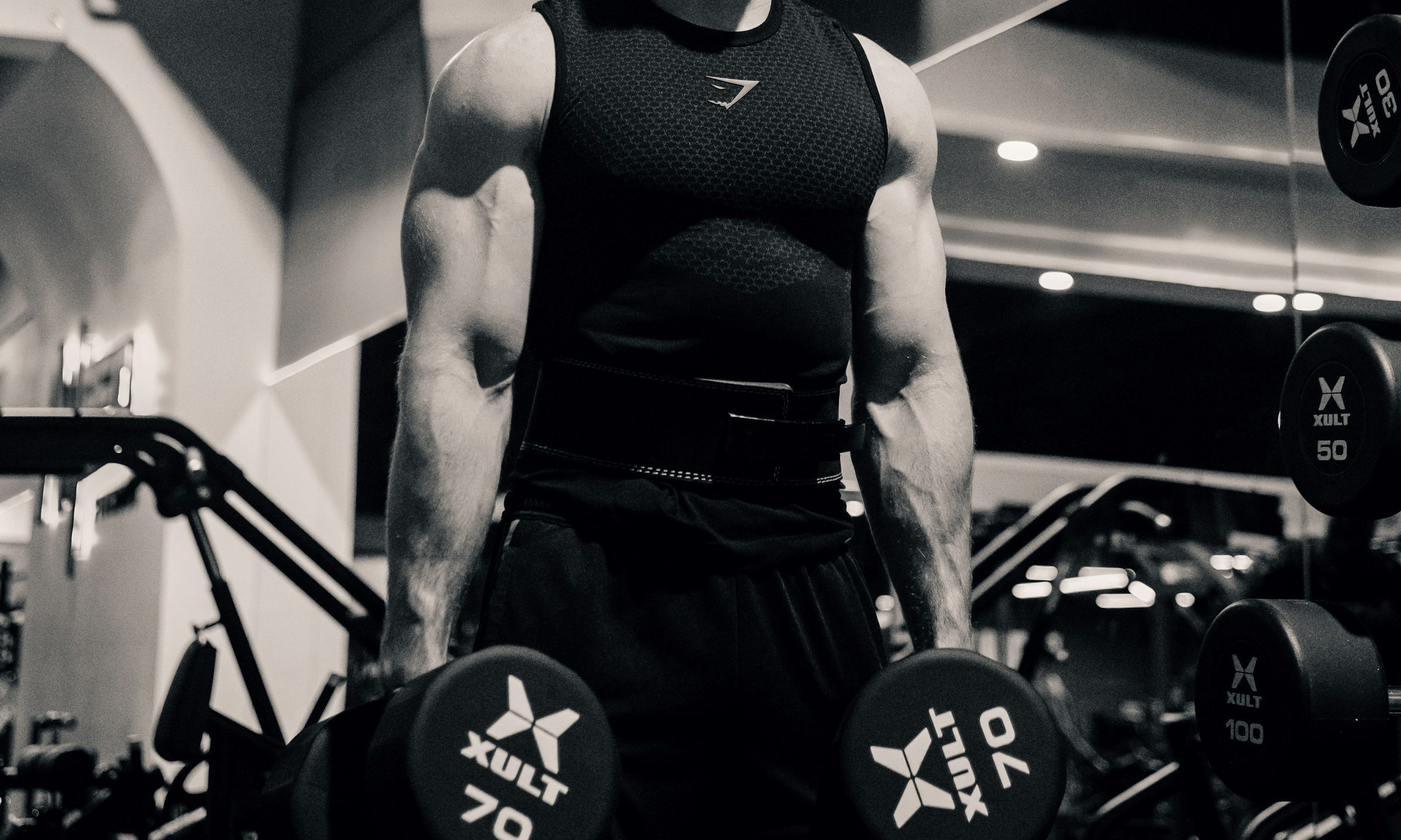 Man in gym black and white pic holding dumbbells down at his sides ready to do dumbbell squat exercise in gym