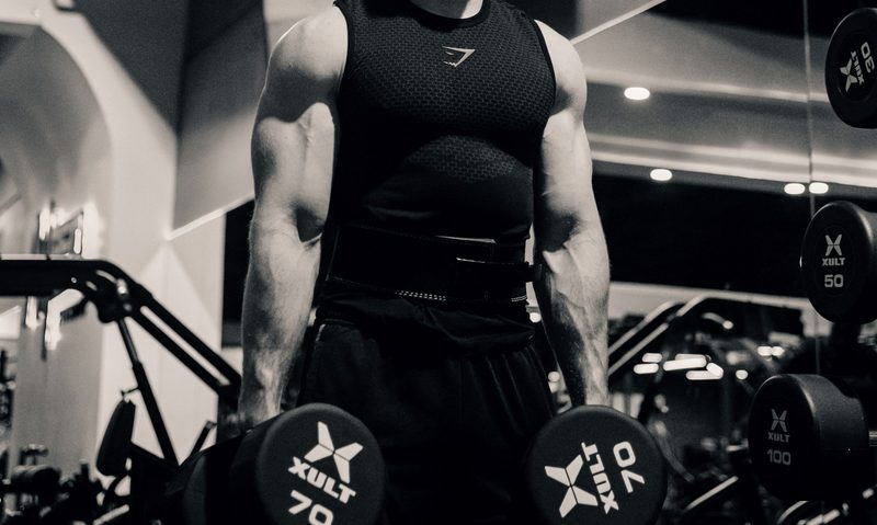 man in gym black and white pic holding dumbbells down at his sides ready to do dumbbell squat exercise in gym
