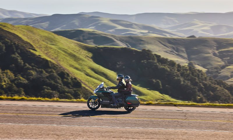 Two people riding a Honda Gold Wing on a mountain road with mountains and valleys in the background.