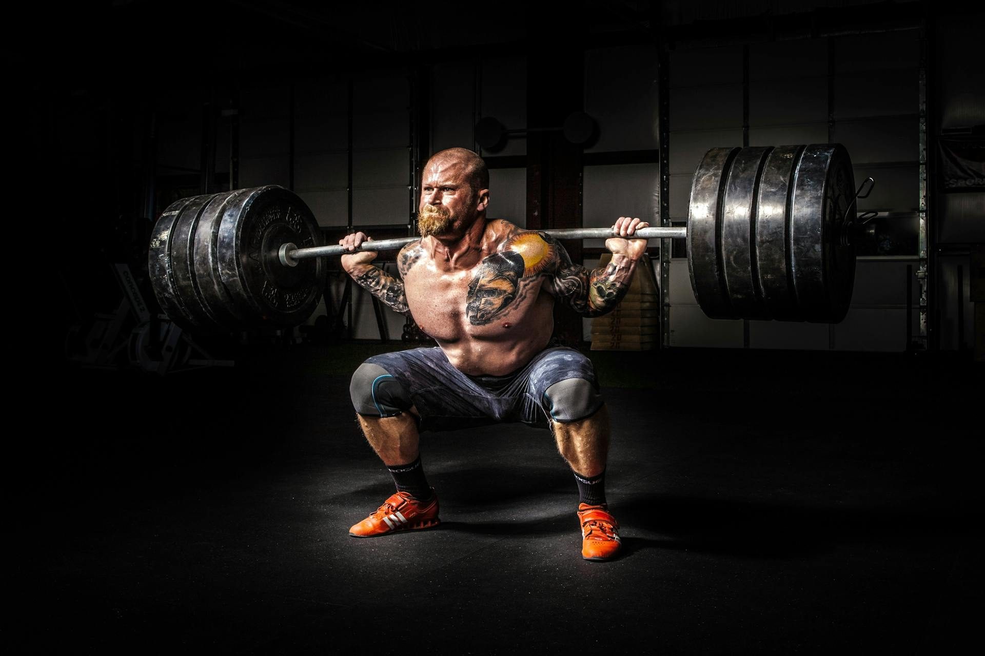 man performing barbell back squat shirtless tattoo dark backround heavy weight