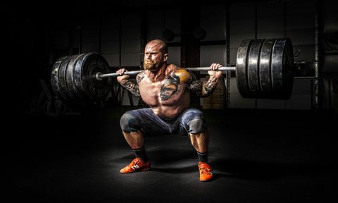 man performing barbell back squat shirtless tattoo dark backround heavy weight