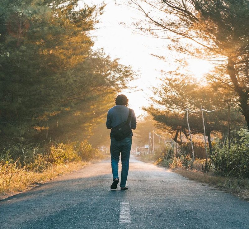 man walking outdoors through sunlit road trees
