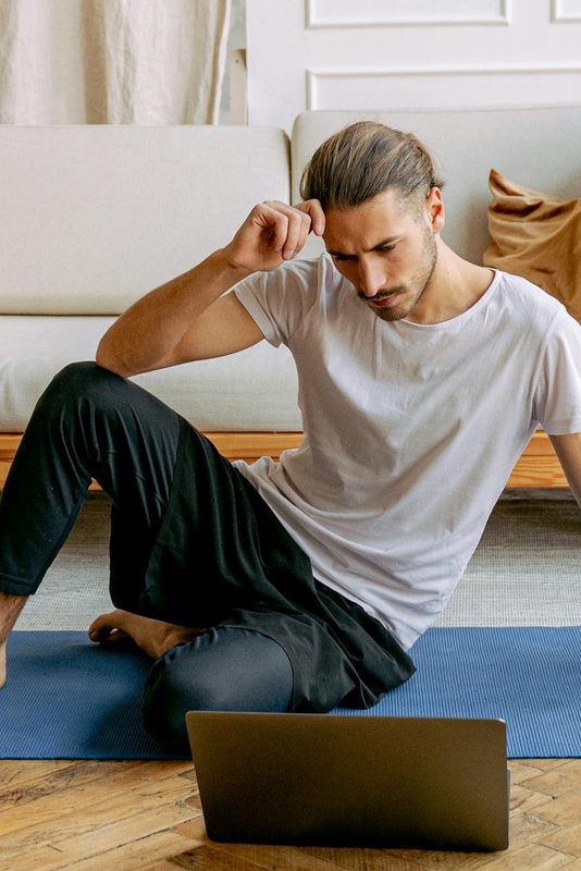 man sitting on yoga mat at home workout laptop exercise class thinking