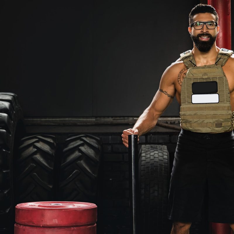 man wearing weighted vest and glasses smiling in gym next to tires