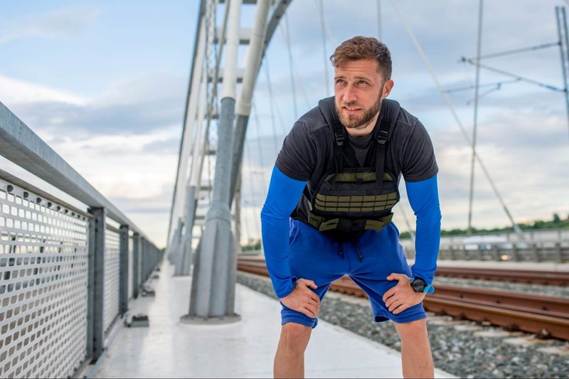 man taking break from running working out walking wearing weighted vest outdoors in city near train track
