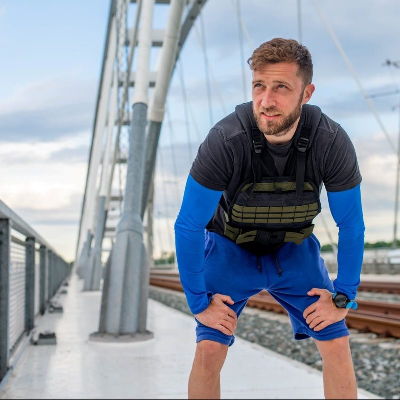 Man taking break from running working out walking wearing weighted vest outdoors in city near train track