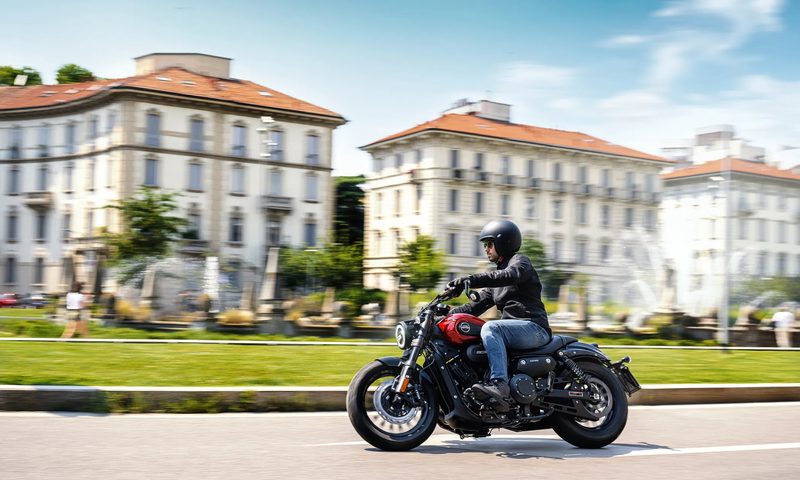 Person riding a 2025 Benda Chinchilla 300 on a city street with large residence buildings in the background.