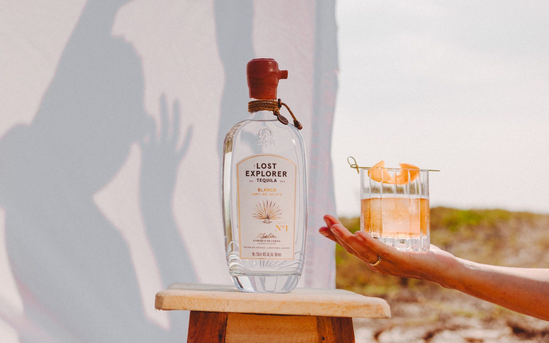 A hand holding out an old fashioned in front of a bedsheet