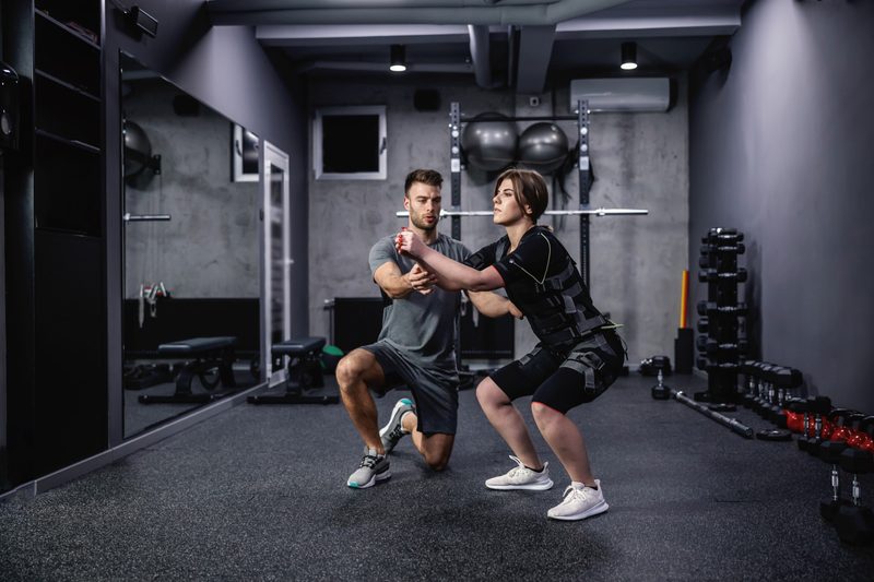 man helping woman do squats wearing EMS suit exercise electrical muscle stimulation
