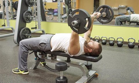 Man doing skull crushers with an EZ curl barbell on bench gym