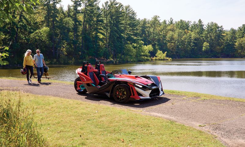 A couple with helmets walking toward a Polaris Slingshot 10 anniversary limited edition parked on a path on the side of a lake.