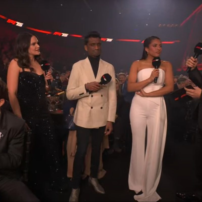 F1 75 Live - Host Jack Whitehall chatting with F1 TV commentators Laura White, Baretto, and Ariana Bravo near Ferrari driver Charles Leclerc's table.