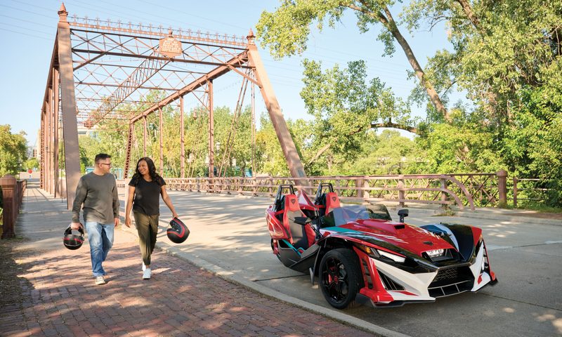 Two people carrying helmets walking to a Polaris Slingshot 10 anniversary limited edition parked on the side of the road.