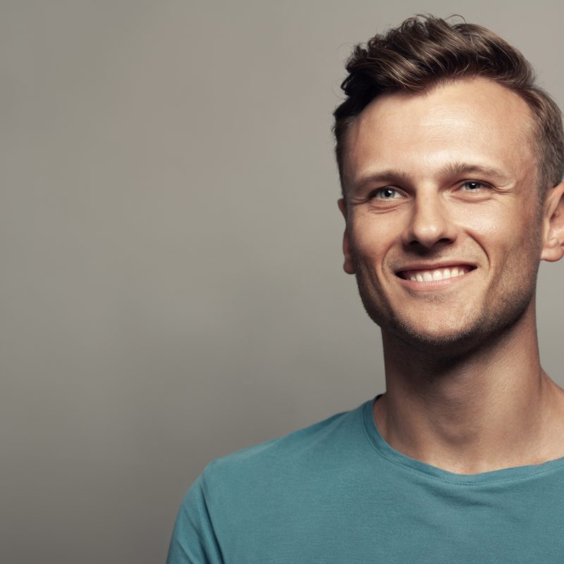Male beauty concept. Portrait of smiling 30-year-old man standing in blue t-shirt over gray background . Close up. Classic style. Wavy glossy blond hair. Studio shot