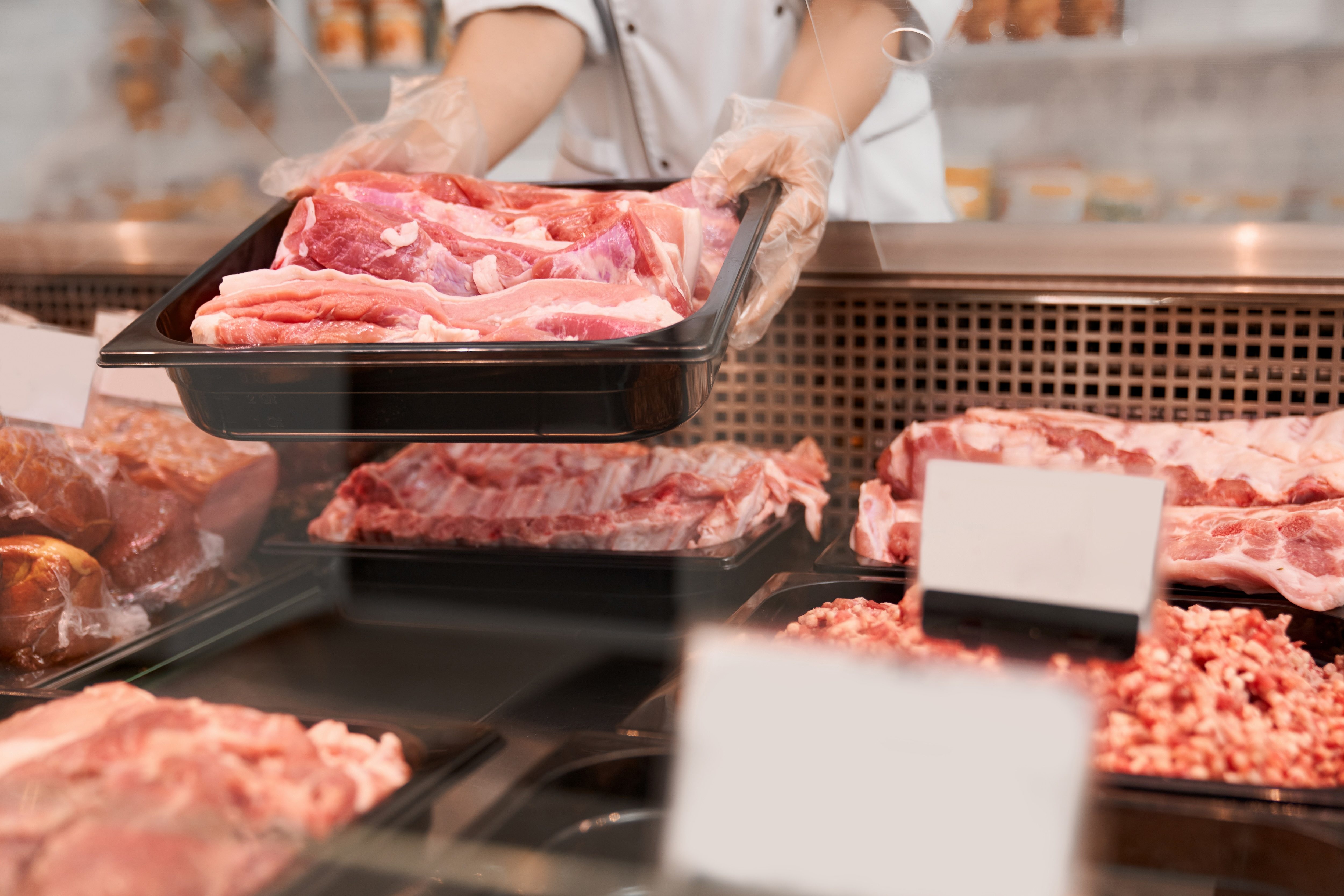 Close up of raw meat pieces in refrigerator with price tags ready for sale in meat department of store. Female hands putting out of glass counter plate with sliced fresh steaks.