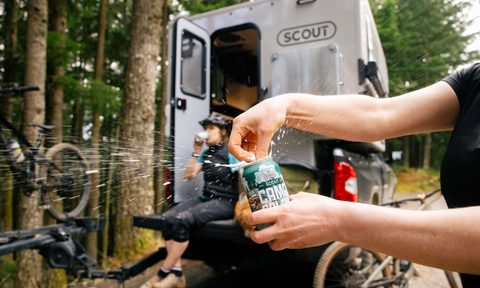 Closeup of hands cracking open a 10 Barrel Camp Coldie beer can in front of a Scout Campers truck camper.