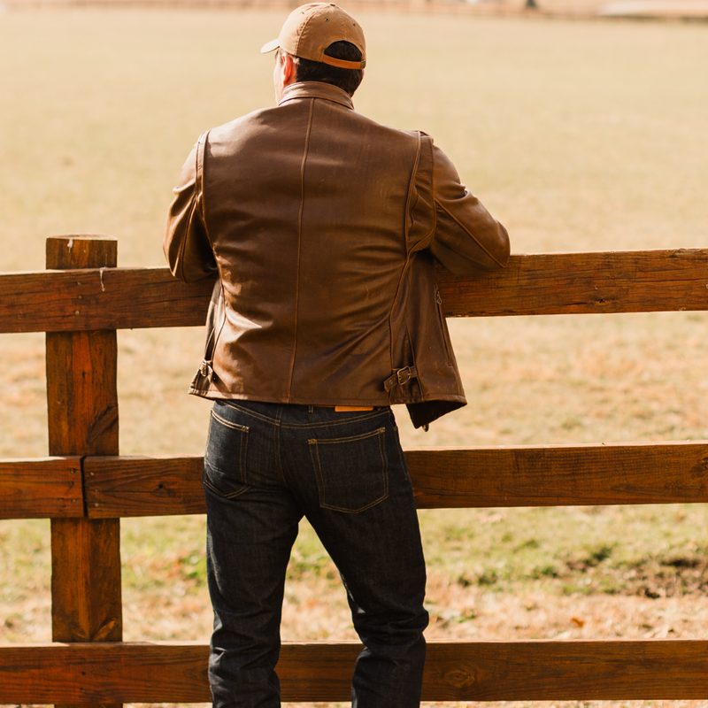 A man leaning on a post fence facing away from the camer wearing a Ball and Buck x Schott NYC Leather Jacket.
