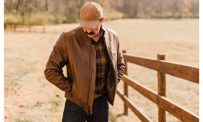 A man standing next to a post fence wearing a Ball and Buck x Schott NYC Leather Jacket.