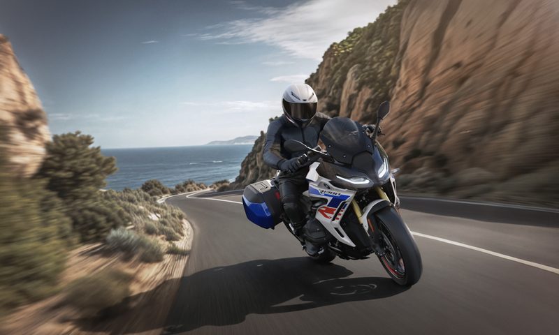 Rider on a 2026 BMW R 1300 RS driving on roadway between high rocks with ocaen in the background.