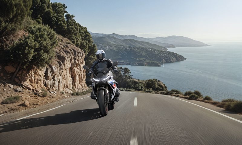 Rider on a 2026 BMW R 1300 RS driving on roadway directly at the viewer with ocean in the background.