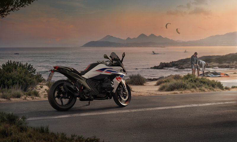 Two people folding up a tent on a small strip of sand with a 2026 BMW R 1300 RS parked roadside in the foreground.