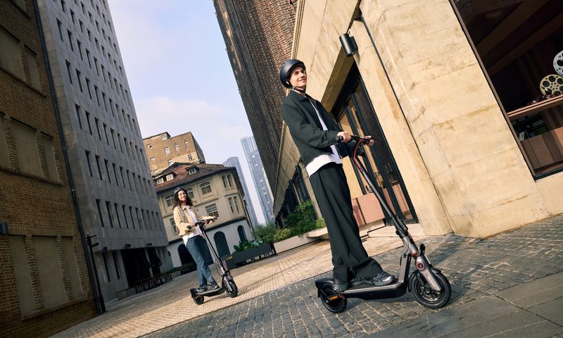 Two people wearing helmets riding Segway Ninebot F3 eKickScooters on stone walkways in city setting.