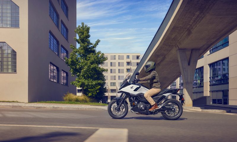 A left profile shot of a rider on a 25 Honda NC750X DCT on a city street with large buildings and a freeway ramp in the background.