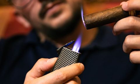 a bespectacled man lighting a cigar using a torch lighter