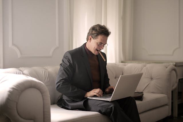 man in black suit sitting on white couch smoking a cigar while using his laptop