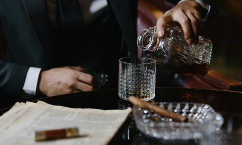 man in black suit pouring drink on glass with cigar in ashtray
