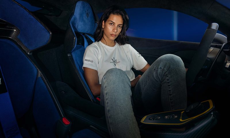 Model sitting in a Maserati driver seat facing the passenger side wearing clothes from the Sparco X Maserati fan collection.