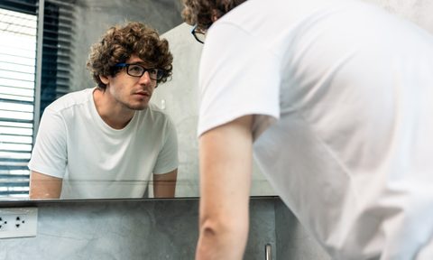 Caucasian young man washing and clean face with facial foam and water. Attractive male washing face for healthy beauty treatments and skin care then looking at mirror and wear eyeglasses in bathroom