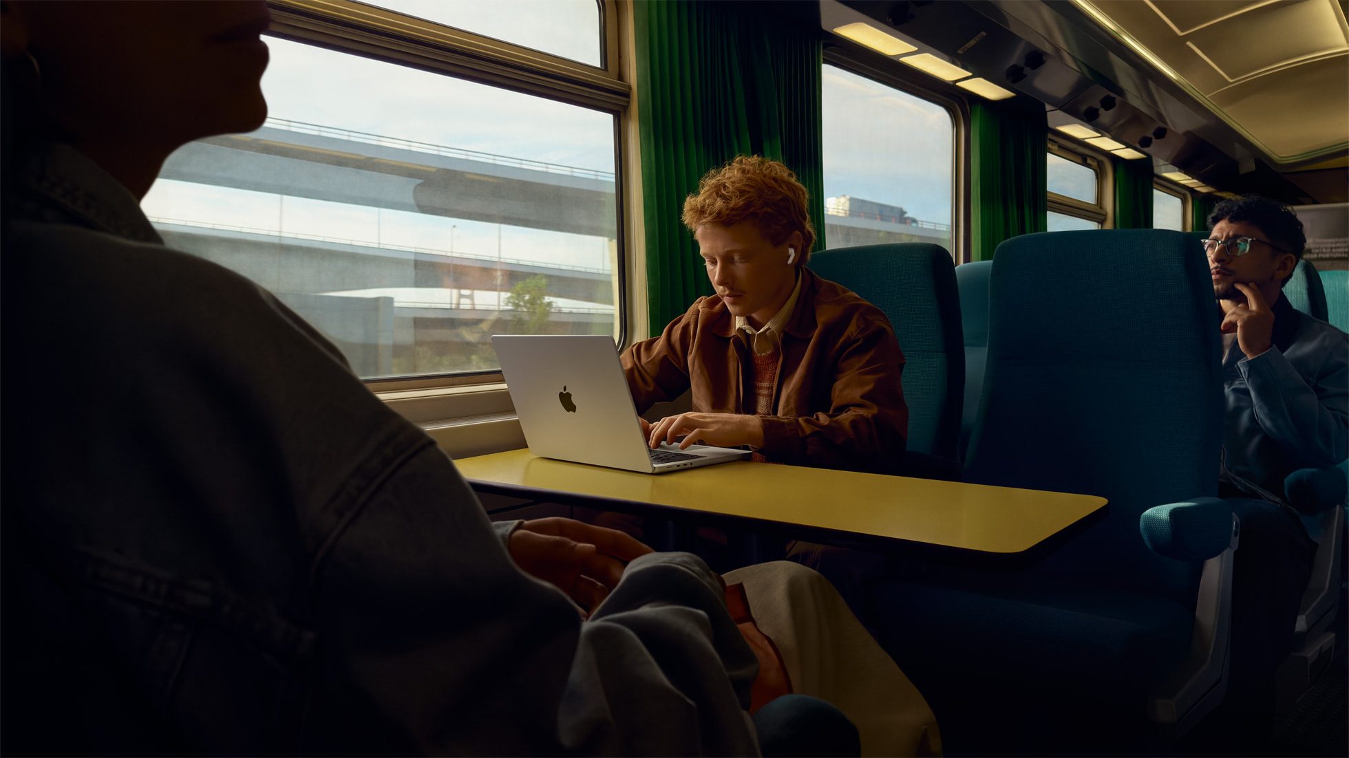 Man on a train working on a MacBook while wearing AirPods Pro 3 earbuds