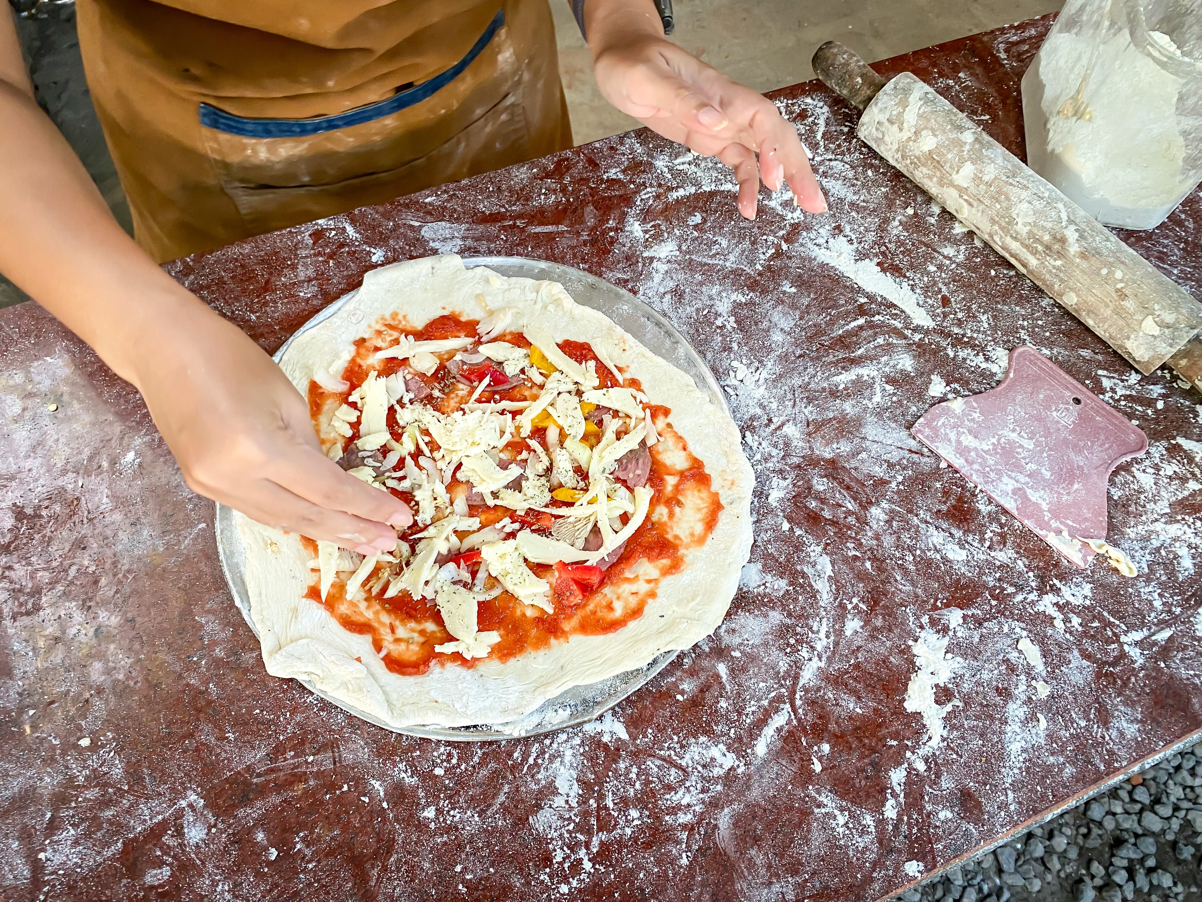 A woman wearing apron is sprinkling the dough with mozzarella cheese, sausage, peppers and vegetables toppings during a pizza cooking class session